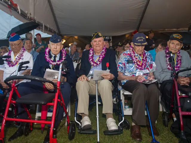 The image shows a group of veterans sitting in wheelchairs under a tent, wearing garlands and caps....