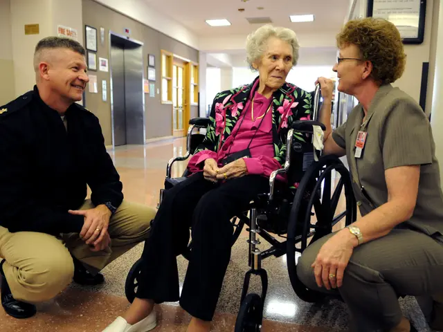 The image shows an elderly woman in a wheelchair talking to two nurses in a hospital hallway. The...