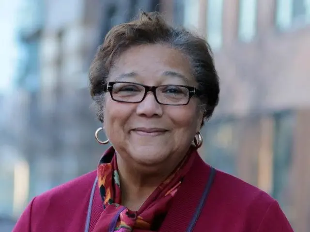 The image shows a woman wearing glasses and a scarf standing in front of a building, with a blurred...