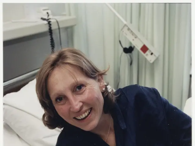 The image shows a woman in a hospital bed, cradling a baby in her arms. She is wearing a blue dress...