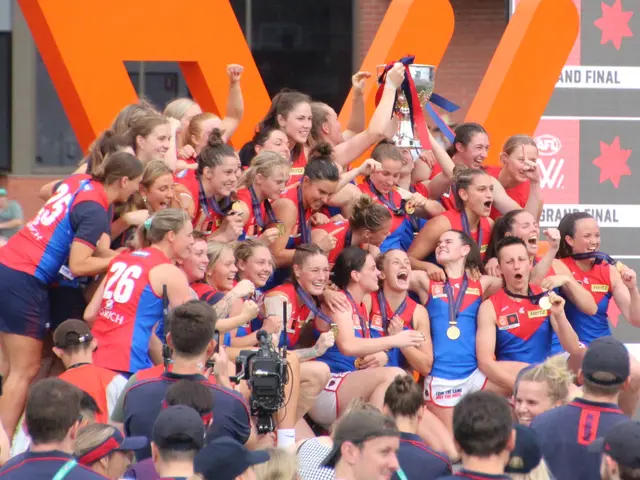 The image shows a group of women's AFL players celebrating with a trophy in front of a cheering...