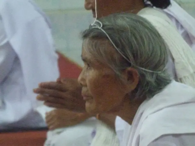 The image shows an elderly woman laying in a hospital bed with her hands clasped together,...