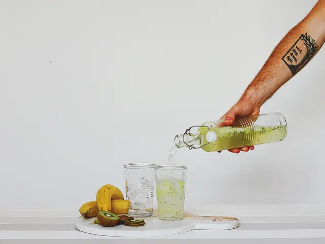 The image shows a person pouring a green smoothie from a bottle into a glass on top of a white...