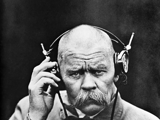 The image shows an old man with a mustache wearing headphones and talking on a cell phone. He is...