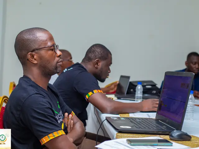 The image shows a group of men sitting around a table with laptops, water bottles, mobile phones,...