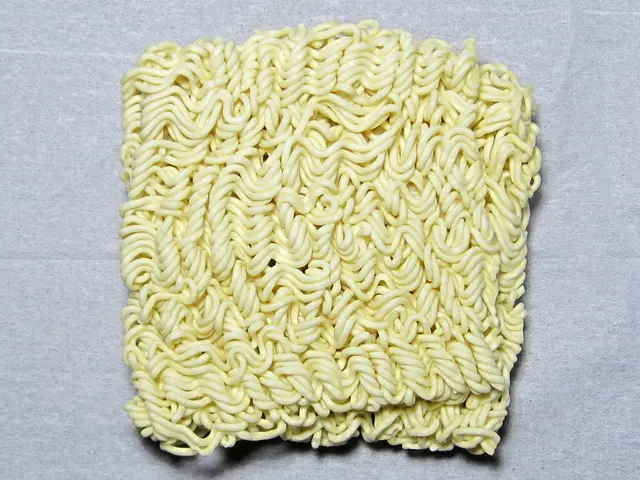 The image shows a pile of instant noodles sitting on top of a table. The noodles are a light yellow...