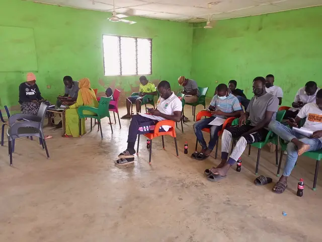 The image shows a group of people sitting in chairs in a room with a wall in the background, a...