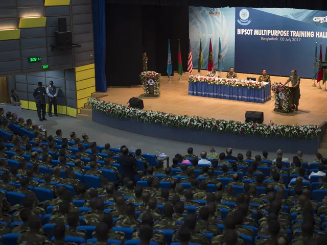 The image shows a large group of people sitting in chairs in front of a stage, with a few people...