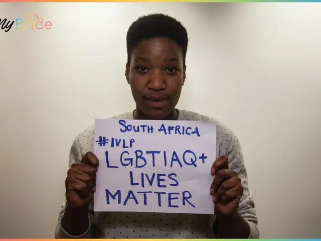 The image shows a woman standing in front of a wall, holding up a sign that reads "LGBTQ+ Lives...