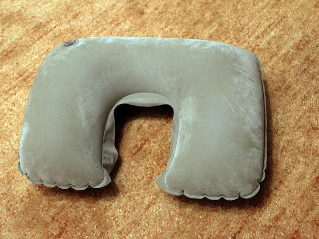 The image shows a gray neck pillow sitting on top of a brown carpeted floor. The pillow is made of...