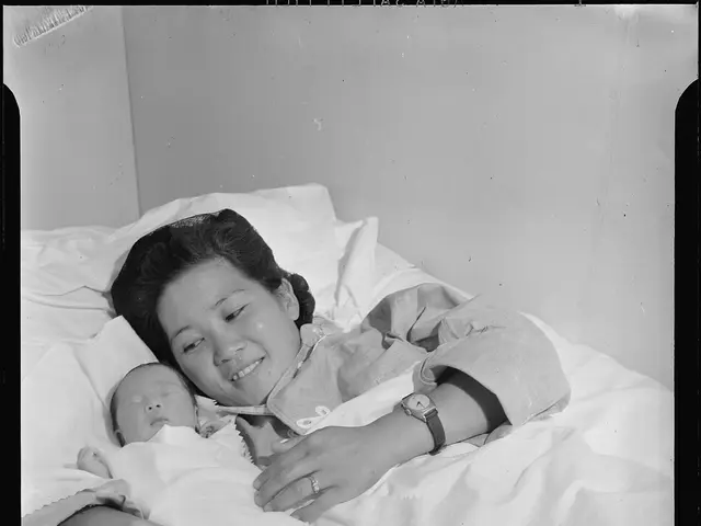 The image shows a woman laying in a hospital bed, cradling a baby in her arms. She is wearing a...