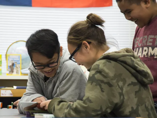 The image shows three students sitting at a table in a classroom, looking at a book. On the table...