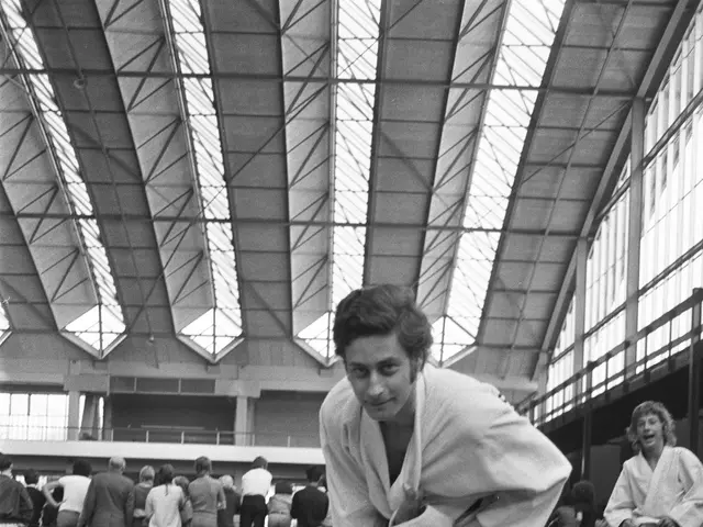 The image shows a black and white photo of two people doing judo in a gym, surrounded by a group of...