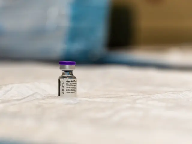 The image shows a small vial of vaccine sitting on top of a white sheet, with something written on...
