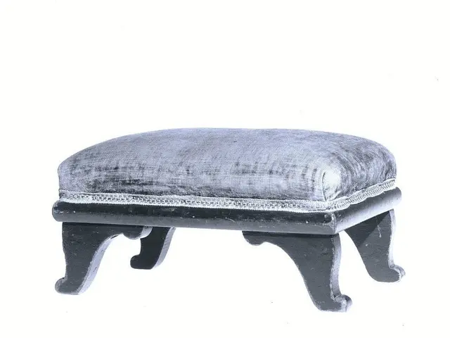 The image shows a small black footstool with a cushion on top of it, set against a white...