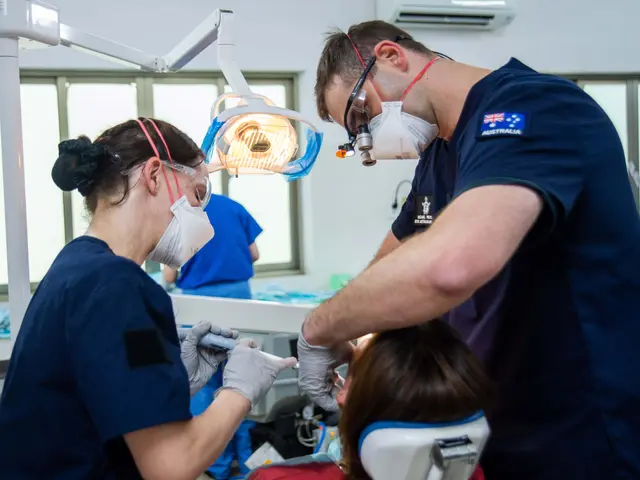 The image shows two people in scrubs and masks working on a patient in a dentist's chair. The...