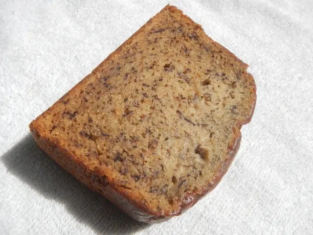The image shows a freshly-baked loaf of banana bread sitting atop a white towel. The golden-brown...