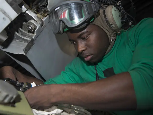 The image shows a man wearing a green t-shirt, headphones, and goggles, working on a machine in a...