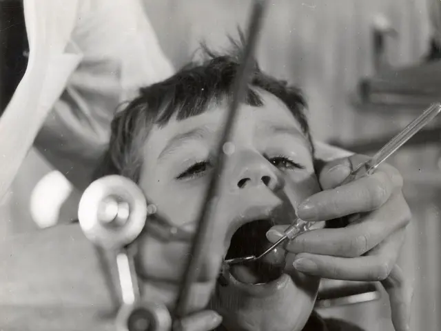 The image shows a black and white photo of a young boy getting his teeth checked by a dentist. The...