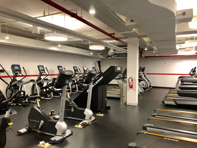 The image shows a gym with a row of treadmills and elliptical machines on the floor, a pillar in...