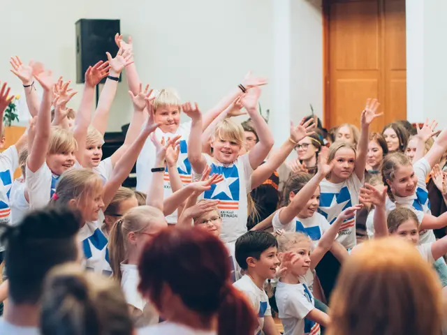 The image shows a group of children standing in front of a crowd with their hands in the air,...