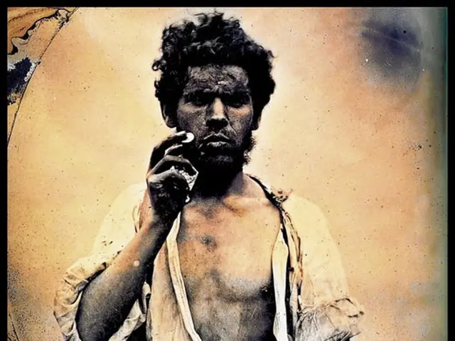 The image shows a man sitting and smoking a cigarette. He is wearing a white shirt and has a...