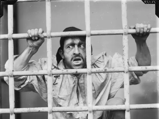 The image shows a black and white photo of a man sitting behind bars in a jail cell. He is gripping...
