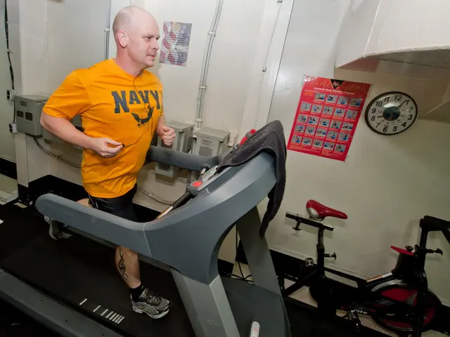 The image shows a man running on a treadmill in a gym. He is wearing a yellow t-shirt and black...