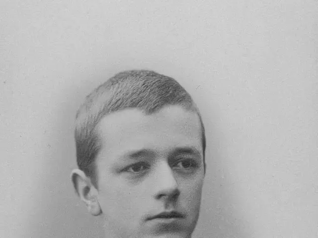 The image shows a black and white photo of a young man wearing a suit. His face is clearly visible,...