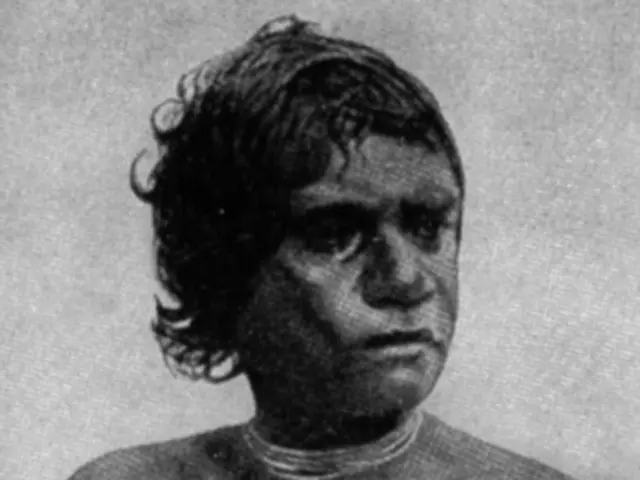 The image shows a black and white photo of a man with a tattoo on his chest. At the bottom of the...