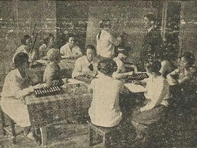 The image shows an old black and white photo of a group of people sitting around a table playing...