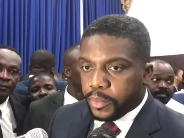 The image shows a man in a suit and tie speaking into a microphone in front of a group of people,...