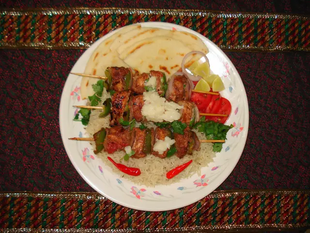 The image shows a plate of kebabs with rice and vegetables on a table. The plate is filled with a...