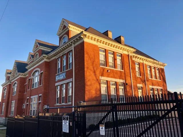 The image shows a large brick building with a black fence in front of it, surrounded by lush green...