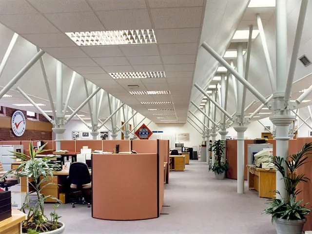 The image shows an empty office with lots of desks and chairs, houseplants, pillars, a clock on the...