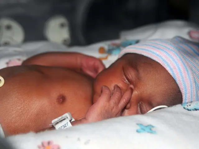 The image shows a premature baby sleeping peacefully in a hospital bed, wearing a cap and...