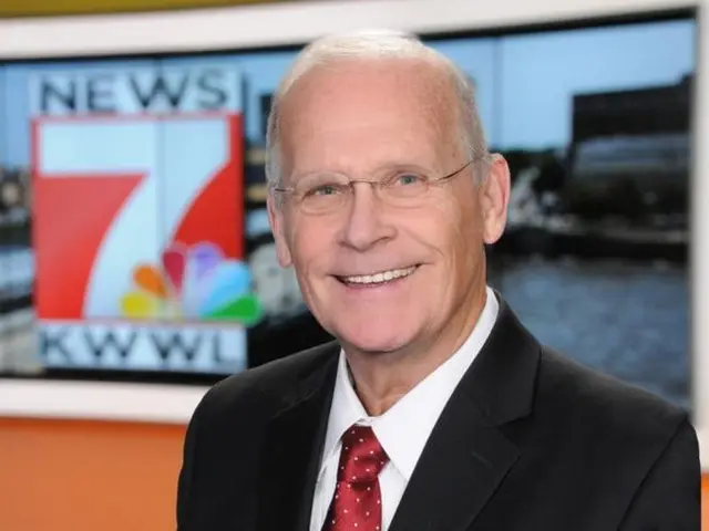 The image shows a man in a suit and tie standing in front of a TV, smiling. He is wearing...