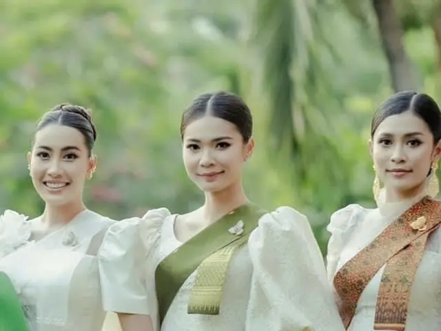 The image shows three women standing next to each other in traditional Thai clothing, smiling and...