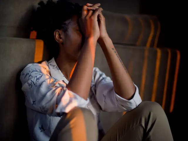 The image shows a woman sitting on a couch with her head in her hands, her expression conveying a...