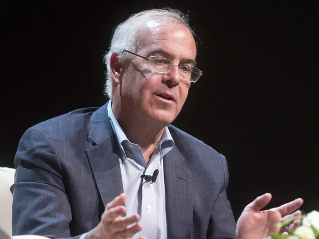 The image shows a man in a suit and tie sitting on a couch, wearing glasses and a microphone on his...