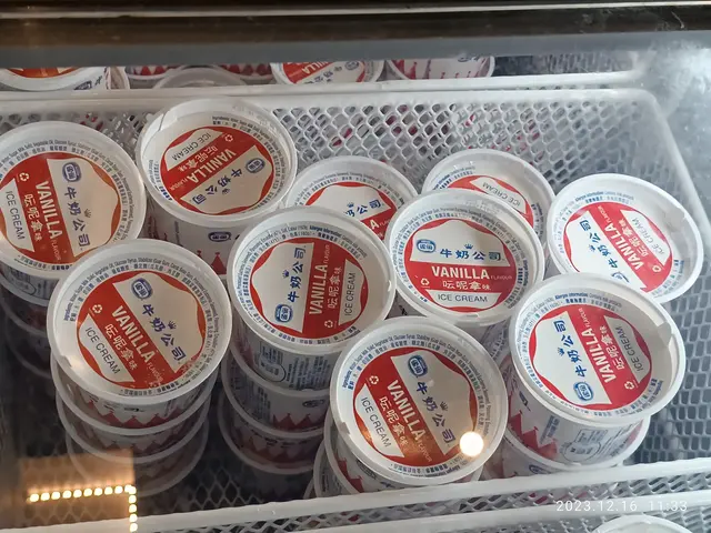 The image shows a display case filled with lots of different types of ice cream, each with a white...